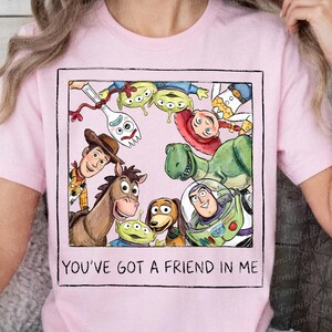 May include: A light pink t-shirt featuring a square graphic of various animated characters including a cowboy, a space ranger, a dinosaur, a horse, a spork, and aliens. The text "YOU'VE GOT A FRIEND IN ME" is printed in black below the graphic.