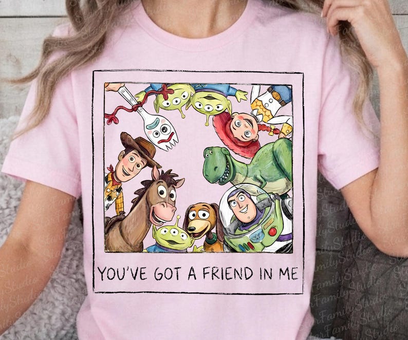 May include: A light pink t-shirt featuring a square graphic of various animated characters including a cowboy, a space ranger, a dinosaur, a horse, a spork, and aliens. The text "YOU'VE GOT A FRIEND IN ME" is printed in black below the graphic.