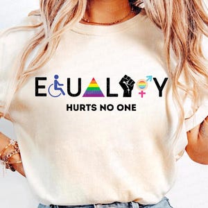 Op de afbeelding: Crèmekleurige sweatshirt met het woord "EQUALITY" in zwarte letters, met symbolen voor handicap, LGBTQ+ en een vuist. Daaronder staat de zin "HURTS NO ONE".