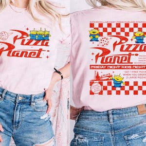 K&ouml;nnte beinhalten: Pinkes T-Shirt mit einer roten und wei&szlig;en Grafik des Pizza Planet-Logos aus dem Toy Story-Film-Franchise. Das Logo enth&auml;lt den Text "Pizza Planet" und drei Alien-Charaktere. Die R&uuml;ckseite des Shirts zeigt ein rot-wei&szlig;es Karomuster mit dem Text "Interstellar Fast-Food Fueling Station" und "Pizza Planet" mit einem kleineren Alien-Charakter.