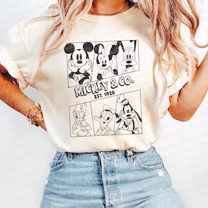 Puede incluir: Una camiseta blanca con un estampado gráfico en blanco y negro de Mickey Mouse, Minnie Mouse, Donald Duck y Goofy. El gráfico incluye el texto "MICKEY & CO. EST. 1928".