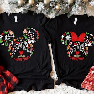 Könnte beinhalten: Zwei schwarze T-Shirts mit einem Weihnachts-Mickey-Mouse- und Minnie-Mouse-Design. Das Design zeigt eine rote Schleife, einen Weihnachtsbaum, einen Schneemann und den Text "Joy to the World" und "Merry Christmas".