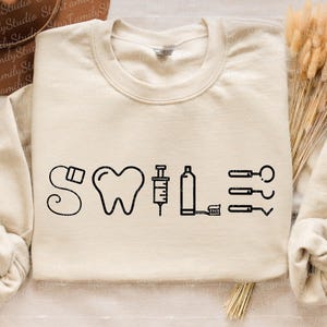 May include: A beige sweatshirt with the word "SWILE" printed in black. The word is made up of dental tools, including a toothbrush, syringe, floss, and a tooth.