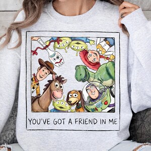 May include: A light gray crewneck sweatshirt featuring a square graphic print of various animated characters including a cowboy, a space ranger, a dinosaur, a horse, and aliens. The text "YOU'VE GOT A FRIEND IN ME" is printed below the illustration.