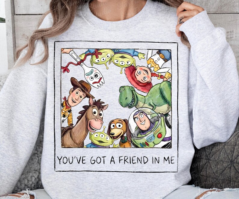May include: A light gray crewneck sweatshirt featuring a square graphic print of various animated characters including a cowboy, a space ranger, a dinosaur, a horse, and aliens. The text "YOU'VE GOT A FRIEND IN ME" is printed below the illustration.