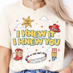 Może przedstawiać: Kremowy t-shirt z napisem "I KNEW IT I KNEW YOU" w kolorze ż&oacute;łtym z niebieskim obrysem. Grafiki przedstawiają złotą gwiazdę szeryfa, czerwony kowbojski kapelusz, czerwone kowbojki, bransoletkę z koralik&oacute;w z napisem "You've got a friend in me" oraz klocki.