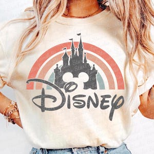 Pode incluir: Uma camiseta branca com um design de arco-íris retrô que apresenta uma silhueta do castelo de Cinderela e a palavra "Disney" em uma fonte estilizada.