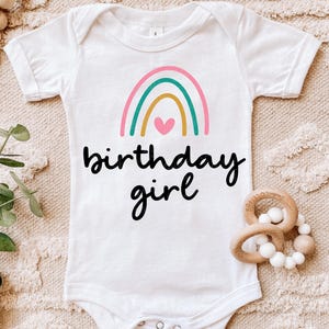 Könnte beinhalten: Weißer Baby-Body mit einem bunten Regenbogen-Motiv und dem Schriftzug "birthday girl" in schwarzer Schrift. Der Regenbogen hat rosa, türkis und goldfarbene Bögen mit einem rosa Herz. Ein süßes Outfit für den Geburtstag eines Babys.