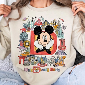May include: A beige sweatshirt with a vintage-style graphic of Mickey Mouse in front of a colorful illustration of Walt Disney World. The text "Walt Disney World" is printed below the graphic.