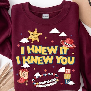 Może przedstawiać: Bordowa bluza z okrągłym dekoltem z nadrukiem graficznym z napisem I KNEW IT I KNEW YOU. Wz&oacute;r zawiera ż&oacute;łtą gwiazdę szeryfa, czerwony kowbojski kapelusz, czerwone kowbojki, bransoletkę z koralik&oacute;w z tekstem oraz stos klock&oacute;w z literami.