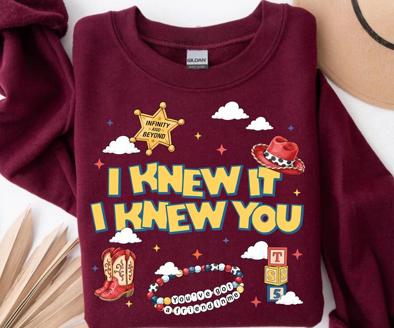 Może przedstawiać: Bordowa bluza z okrągłym dekoltem z nadrukiem graficznym z napisem I KNEW IT I KNEW YOU. Wz&oacute;r zawiera ż&oacute;łtą gwiazdę szeryfa, czerwony kowbojski kapelusz, czerwone kowbojki, bransoletkę z koralik&oacute;w z tekstem oraz stos klock&oacute;w z literami.