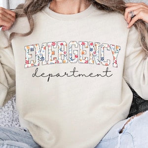 K&ouml;nnte beinhalten: Ein cremefarbener Pullover mit dem Text "EMERGENCY department" in schwarzen Buchstaben mit einem Blumenmuster.