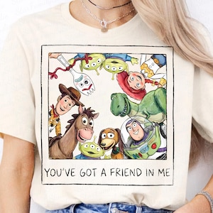 May include: A cream-colored t-shirt featuring a square graphic print of Toy Story characters including Woody, Buzz Lightyear, Jessie, Rex, Bullseye, Forky, and aliens. The text below the illustration reads, "YOU'VE GOT A FRIEND IN ME."