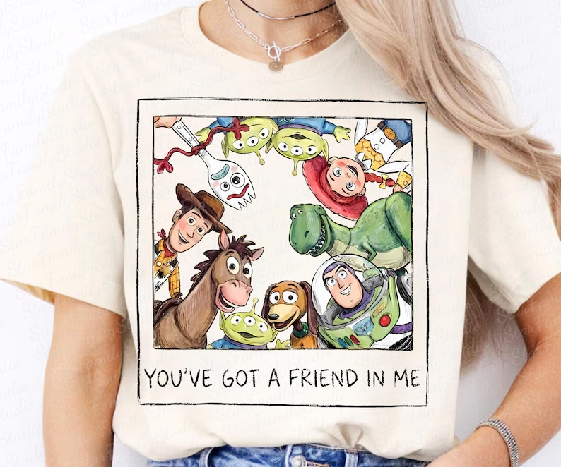 May include: A cream-colored t-shirt featuring a square graphic print of Toy Story characters including Woody, Buzz Lightyear, Jessie, Rex, Bullseye, Forky, and aliens. The text below the illustration reads, "YOU'VE GOT A FRIEND IN ME."