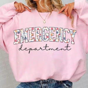 K&ouml;nnte beinhalten: Rosafarbener Pullover mit einem Blumendruck, auf dem "EMERGENCY department" in wei&szlig;en Buchstaben steht.