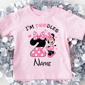 K&ouml;nnte beinhalten: Rosa Kleinkind-T-Shirt mit dem Text "I'm Twoodles Name". Das Design zeigt eine gro&szlig;e Zahl 2 mit wei&szlig;en Punkten, eine Minnie-Maus-Grafik und zwei rosa Schleifen. Das T-Shirt hat kurze &Auml;rmel.