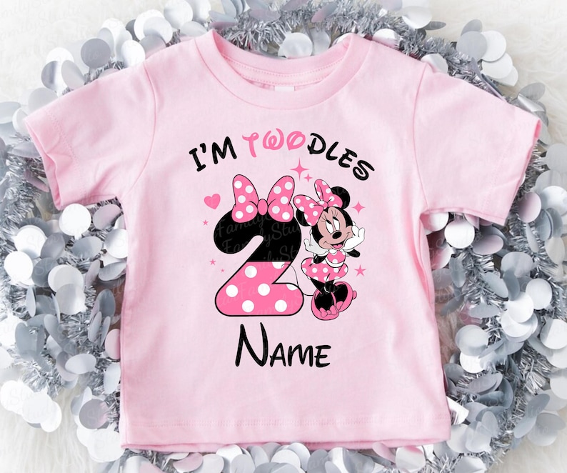 K&ouml;nnte beinhalten: Rosa Kleinkind-T-Shirt mit dem Text "I'm Twoodles Name". Das Design zeigt eine gro&szlig;e Zahl 2 mit wei&szlig;en Punkten, eine Minnie-Maus-Grafik und zwei rosa Schleifen. Das T-Shirt hat kurze &Auml;rmel.