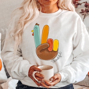 May include: White sweatshirt featuring a turkey hand gesture with the word "Thankful" in script. The turkey's feathers are blue, orange, red, and yellow. The hand is brown. A mug is held in the hands.