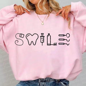 May include: Pink sweatshirt with a black graphic design that says "SWILE" with dental tools and a tooth.