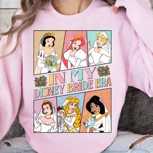Peut inclure: Un sweat-shirt rose avec un imprim&eacute; graphique de princesses Disney en robes de mari&eacute;e. Le texte "In My Disney Bride Era" est imprim&eacute; sur le sweat-shirt.