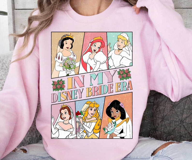 Peut inclure: Un sweat-shirt rose avec un imprim&eacute; graphique de princesses Disney en robes de mari&eacute;e. Le texte "In My Disney Bride Era" est imprim&eacute; sur le sweat-shirt.