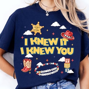 Może przedstawiać: Granatowy t-shirt z ż&oacute;łtym napisem I KNEW IT I KNEW YOU. Grafika zawiera złotą odznakę szeryfa z napisem INFINITY AND BEYOND, czerwony kowbojski kapelusz, brązowe kowbojki, bransoletkę z koralik&oacute;w z napisem YOU'VE GOT A FRIEND IN ME oraz stos trzech klock&oacute;w z literami T, S i 5, otoczonych białymi chmurami i małymi gwiazdkami.