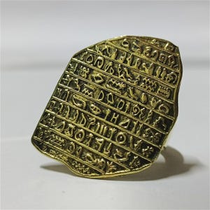 May include: A gold-colored ring featuring a rectangular, textured face with engraved hieroglyphic symbols. The ring has a vintage or antique aesthetic, with a slightly tarnished appearance. The design is reminiscent of ancient Egyptian artifacts.