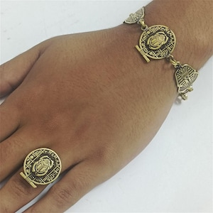May include: A gold-colored ring and bracelet set with an Egyptian theme. The ring features a circular design with a scarab beetle and hieroglyphs. The bracelet has similar circular and stylized pharaoh head charms. The jewelry is worn on a hand.