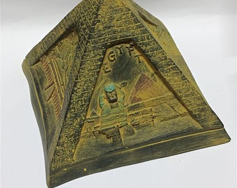 Könnte beinhalten: Ein dekoratives Objekt in Pyramidenform mit einem gealterten, verwitterten Aussehen. Die Pyramide ist hauptsächlich beige und dunkelgrün, mit dem Wort "EGYPT" auf der Vorderseite. Das Design beinhaltet eine Darstellung der Sphinx und anderer ägyptischer architektonischer Elemente.