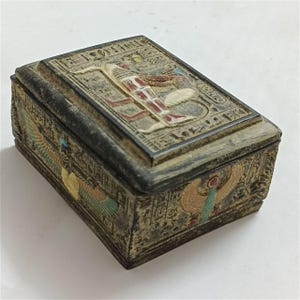 Op de afbeelding: Een rechthoekige doos in antieke stijl met een Egyptisch thema. De doos is voornamelijk donkergrijs met gedetailleerde gravures en hiërogliefen. Het deksel heeft een verhoogd reliëf van een figuur met vleugels en rode en witte accenten. De zijkanten hebben kleurrijke vogel- en vleugelontwerpen.