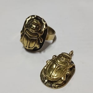 May include: A gold-toned metal ring and a matching pendant, both featuring a three-dimensional scarab beetle design. The ring has a smooth band, while the pendant includes a small loop at the top for attachment, showcasing a polished finish.
