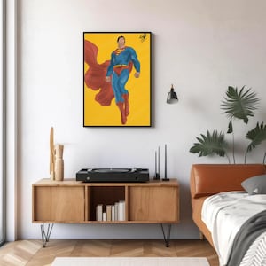 以下が含まれることがあります： ダイナミックなポーズのスーパーマンが描かれた額入りのアートワーク。背景は黄色。スーパーヒーローは、象徴的な青いスーツ、赤いマント、ブーツを着用。白い壁に飾られたアートワーク。