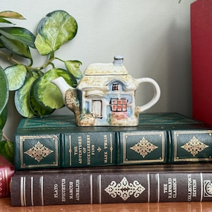 Può includere: Una teiera in ceramica in miniatura a forma di casetta, con tetto di paglia beige, finestre blu e porta bianca. La teiera è posta sopra una pila di libri vintage con copertine verdi e marroni.