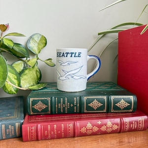 Puede incluir: Una taza de cerámica moteada con la palabra "SEATTLE" en letras azules y dos gaviotas volando. La taza tiene un borde y un asa azules. Descansa sobre una pila de libros antiguos con tapas verdes y rojas.