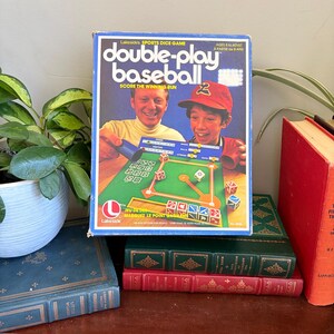 May include: A rectangular board game box titled Double-Play Baseball by Lakeside. The box features a graphic of two people playing a baseball dice game on a green field board with red, white, and blue dice. Text includes Sports Dice Game.
