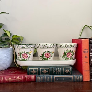May include: Three off-white ceramic flower pots with hand-painted pink flowers and green leaves, set on a matching rectangular tray. Each pot features a green decorative border around the rim. The set is arranged on a stack of hardback books.