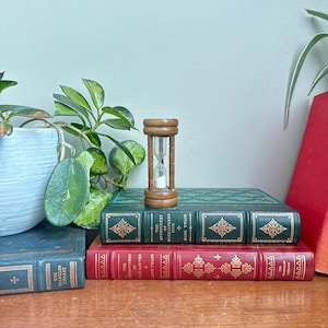 Op de afbeelding: Een houten zandloper met wit zand staat op een stapel vintage boeken. De boeken zijn groen en rood met gouden accenten en titels als "The Adventures of Huckleberry Finn". Een plant in een pot is zichtbaar.