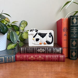 May include: A ceramic cow-shaped butter dish. The dish is white with black spots and a black tail. The cow is lying down on a rectangular base. The dish is on a stack of books with a plant and other books in the background.