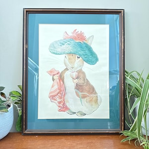 May include: A framed watercolor illustration of a rabbit wearing a teal hat with a pink flower, a brown jacket, and holding a pink cloth and a small pink object. The artwork is set within a dark brown frame with a deep teal matting border.