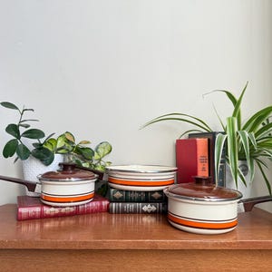 Op de afbeelding: Een set vintage kookgerei met bruine deksels en handvatten, met een crèmekleurige behuizing met oranje en zilveren strepen. De potten staan op een houten oppervlak met boeken en planten. Een klassieke keukengerei set.
