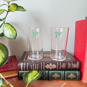 May include: Two clear glass 7 Up glasses with green lettering on a shelf with books. The books are brown, green, and red.