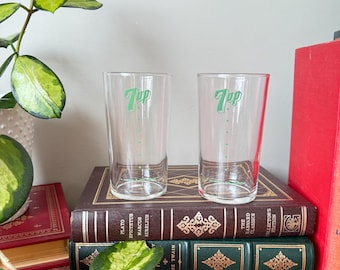 May include: Two clear glass 7 Up glasses with green lettering on a shelf with books. The books are brown, green, and red.