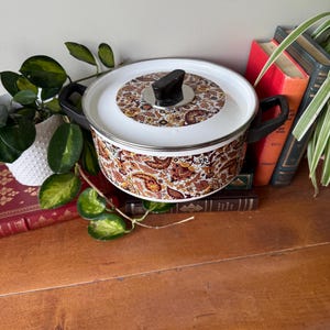 May include: A retro cooking pot with a white lid and black handles. The pot has a brown and orange floral design around the base and on the lid. It is placed on a wooden surface, with books and plants visible in the background.