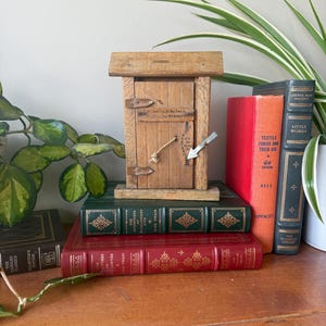 Pode incluir: Uma pequena caixa de madeira em estilo rústico com uma porta com dobradiças e um pequeno telhado, repousando sobre uma pilha de livros antigos. A caixa tem um fecho de metal e um fecho de corda. Os livros são de várias cores.
