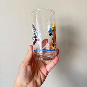 May include: A clear glass tumbler with a blue rim and cartoon graphics. The glass features a design with the word "KEY" and a cartoon character with a ship's wheel. The glass is held by a hand.