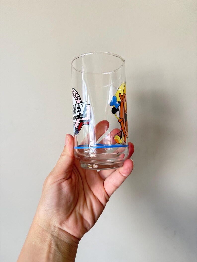May include: A clear glass tumbler with a blue rim and cartoon graphics. The glass features a design with the word "KEY" and a cartoon character with a ship's wheel. The glass is held by a hand.
