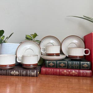 Puede incluir: Un juego de tazas y platillos de café de cerámica vintage. Las tazas y los platillos son de color blanquecino con una banda y un borde marrones. El juego está dispuesto sobre una superficie de madera con libros y una planta en el fondo.