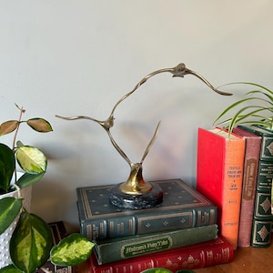 May include: A gold metal sculpture of two birds in flight, perched on a black marble base. The sculpture is displayed on a stack of books with titles including "Andersen's Fairy Tales" and "Textile Fibres and Their Use".