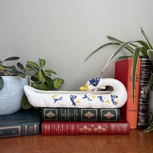 May include: A white ceramic planter shaped like a long boat with a swan head at one end. The exterior features a repeating pattern of small white ducks wearing blue bonnets with white polka dots, accented with yellow beaks and blue details.