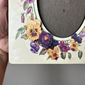 Puede incluir: Un marco de fotos de madera blanca con un borde decorativo de pensamientos morados, amarillos y verdes. El marco tiene una abertura circular para una foto.